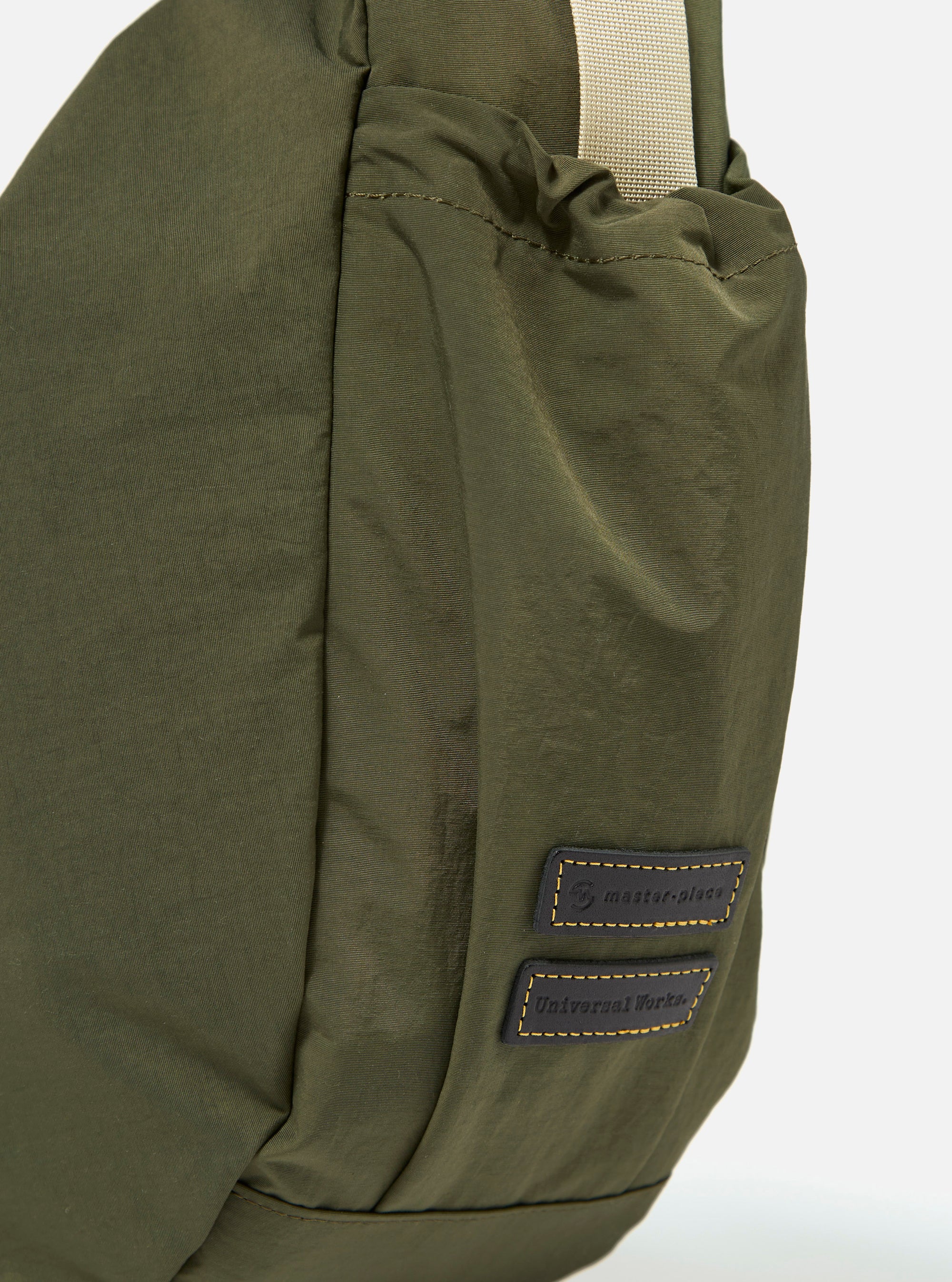 The master-piece x Universal Works Olive Large Canvas Shoulder Bag is shown from the back as a flat lay against a plain white background. The image highlights the rear of the bag, featuring a horizontal webbing strap designed for a suitcase handle and a tonal olive fabric. A small black brand tag is visible near the bottom.