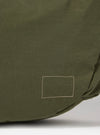 The master-piece x Universal Works Olive Large Canvas Shoulder Bag is shown from the back as a flat lay against a plain white background. The image highlights the rear of the olive green bag, featuring a horizontal webbing strap designed for a suitcase handle and a small black brand tag.