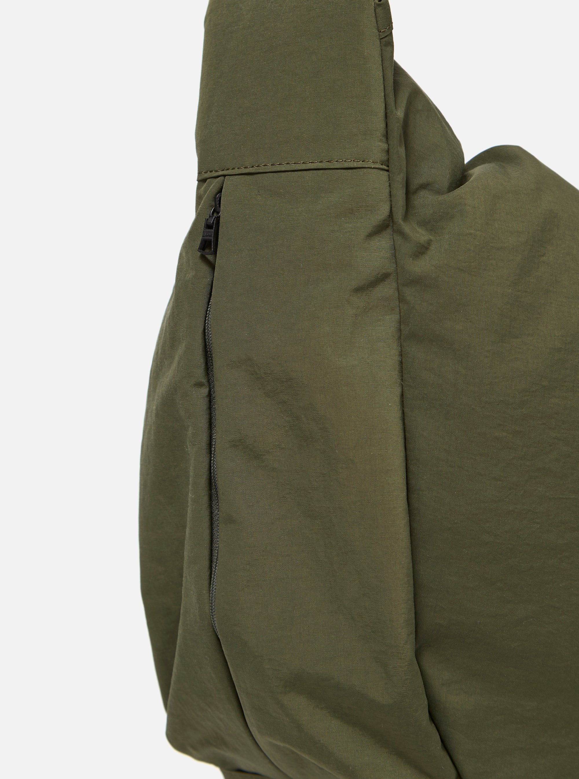 The master-piece x Universal Works Olive Large Canvas Shoulder Bag is shown from the back as a flat lay against a plain white background. The image highlights the bag's olive green canvas construction, featuring a rear compartment with a hook and loop fastener and a webbing strap for attaching to a suitcase handle.