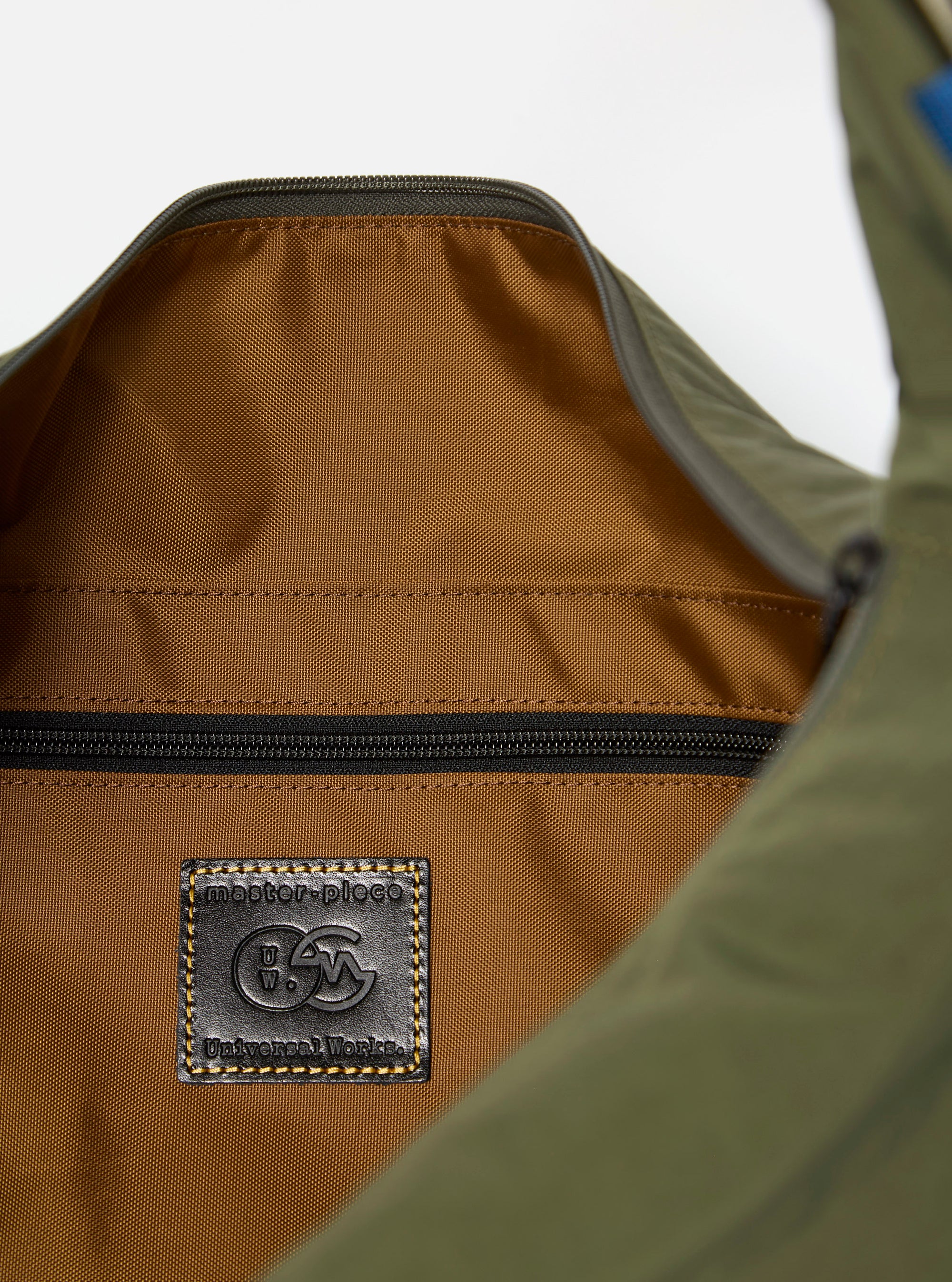 The master-piece x Universal Works Olive Large Canvas Shoulder Bag is shown as a flat lay from a side angle against a plain white background. The bag features an olive green canvas body with a brown leather logo patch, top carry handles, and a detachable olive webbing shoulder strap. The image highlights the bag's depth and the durable texture of the canvas material.