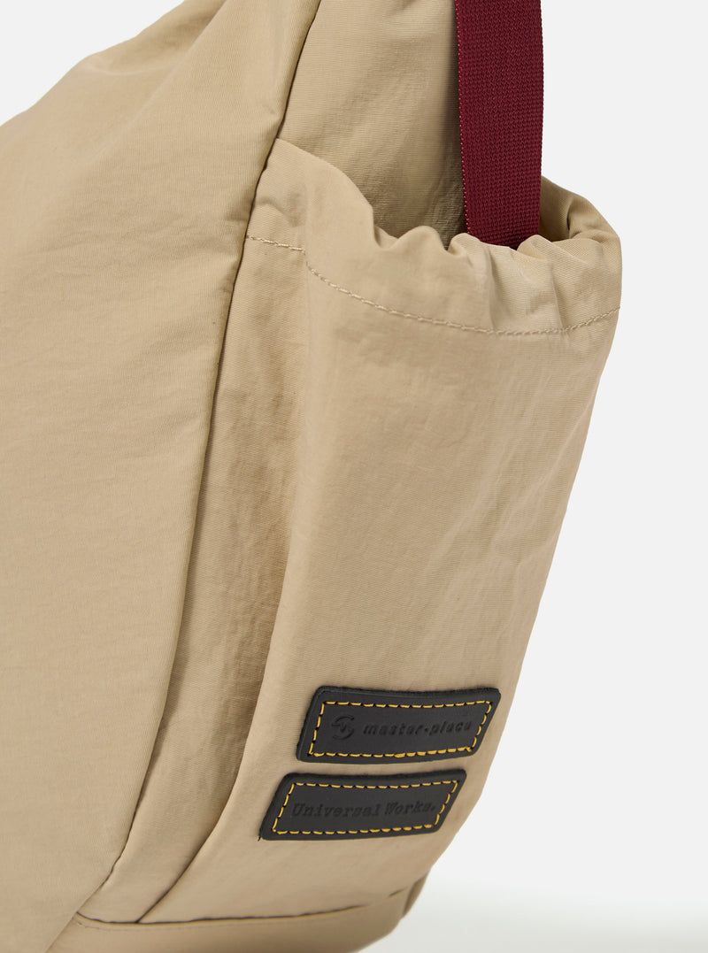 The master-piece x Universal Works Beige Medium Canvas Shoulder Bag is shown from the back as a flat lay against a plain white background. The image highlights the adjustable webbing cross-body strap, a small exterior slip pocket, and the tonal texture of the beige canvas fabric.