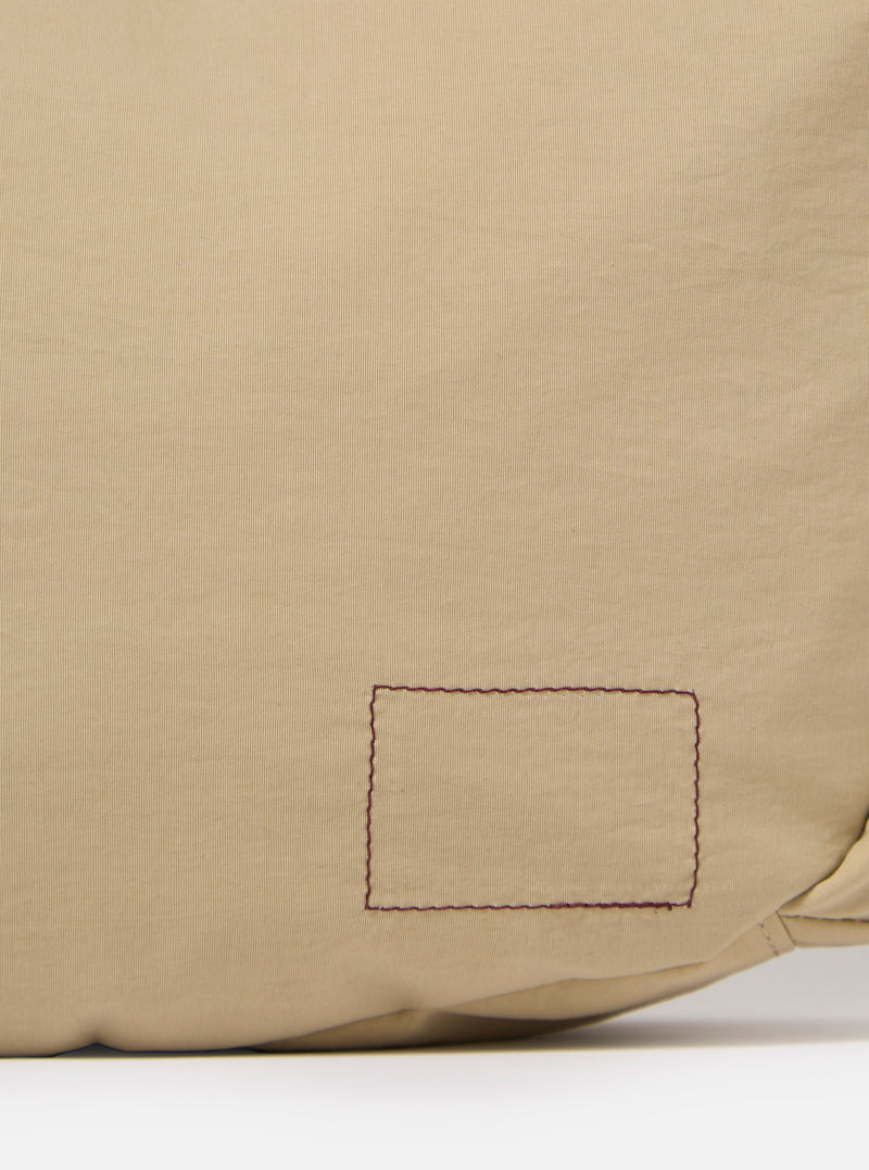 The master-piece x Universal Works Beige Medium Canvas Shoulder Bag is shown from a rear three-quarter angle as a flat lay against a plain white background. The image highlights the adjustable shoulder strap with its black plastic buckle and the clean, structured back panel of the beige canvas bag.