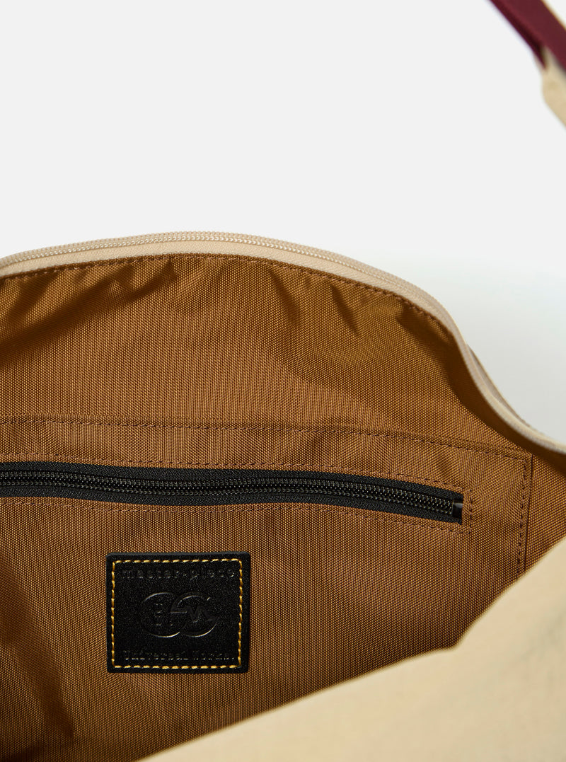The master-piece x Universal Works Beige Medium Canvas Shoulder Bag is shown from the back as a flat lay against a plain white background. The image highlights the adjustable shoulder strap, a small exterior zippered pocket, and a rear sleeve for attaching to a suitcase handle. The bag is made from a durable beige canvas with a visible dual-branded patch.