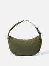The master-piece x Universal Works Olive Medium Canvas Shoulder Bag is shown as a flat lay against a white background. The bag features a rectangular design with a main zippered compartment, a smaller front zippered pocket with a yellow pull tab, and a dual-branded leather logo patch. It includes a detachable and adjustable black webbing shoulder strap.