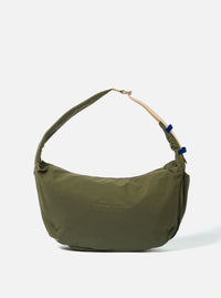 The master-piece x Universal Works Olive Medium Canvas Shoulder Bag is shown as a flat lay against a white background. The bag features a rectangular design with a main zippered compartment, a smaller front zippered pocket with a yellow pull tab, and a dual-branded leather logo patch. It includes a detachable and adjustable black webbing shoulder strap.