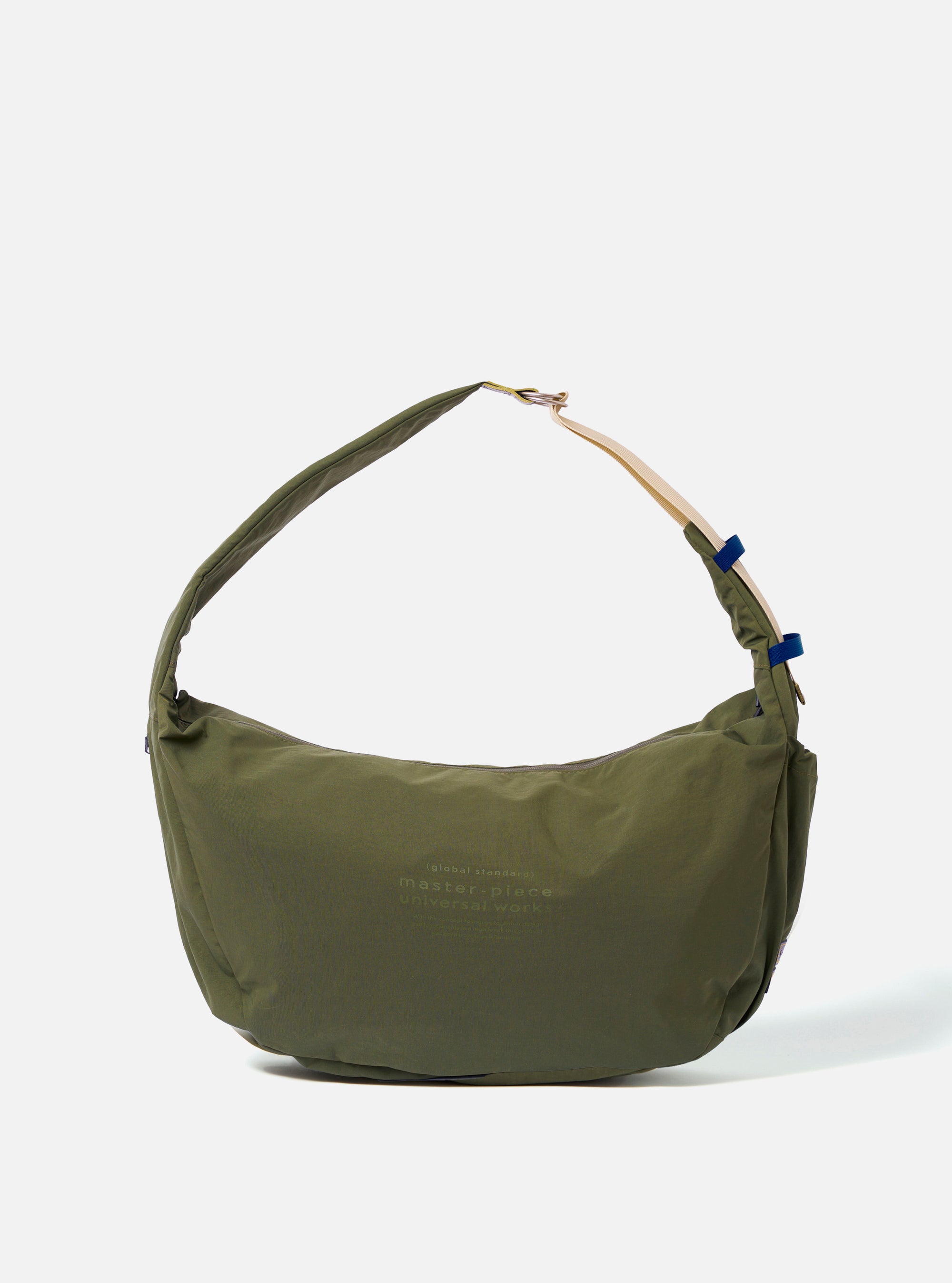 The master-piece x Universal Works Olive Medium Canvas Shoulder Bag is shown as a flat lay against a white background. The bag features a rectangular design with a main zippered compartment, a smaller front zippered pocket with a yellow pull tab, and a dual-branded leather logo patch. It includes a detachable and adjustable black webbing shoulder strap.