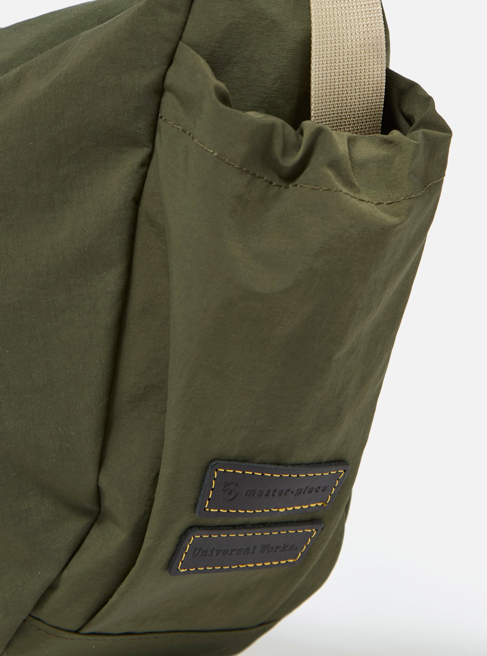 The master-piece x Universal Works Olive Medium Canvas Shoulder Bag is shown from the back as a flat lay against a plain white background. The image highlights the adjustable and detachable olive green webbing strap, the rear pocket with a hook-and-loop closure, and a webbing strap designed for a suitcase handle. The smooth, technical olive green fabric and clean construction are clearly visible.