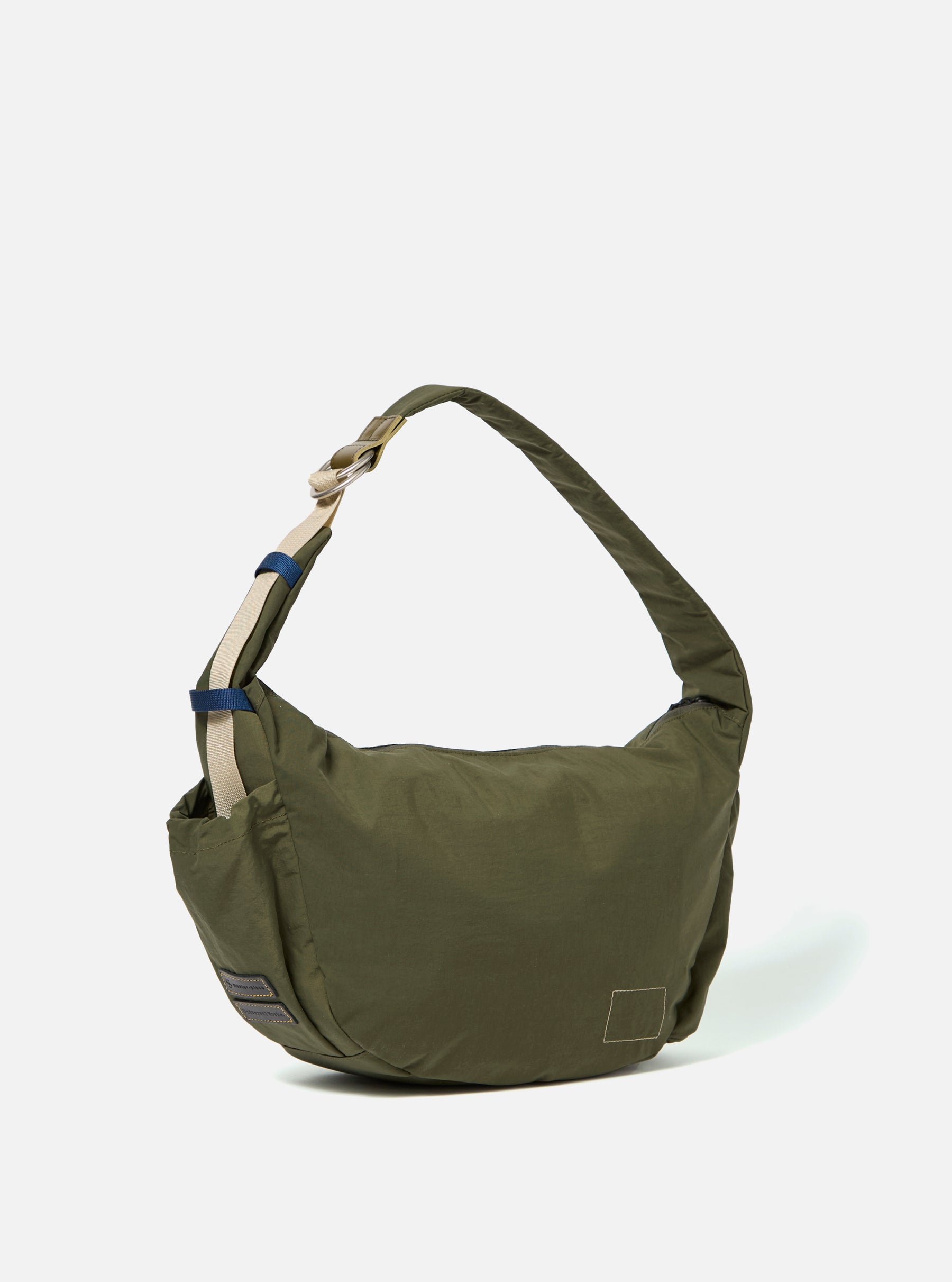 The master-piece x Universal Works Olive Medium Canvas Shoulder Bag is shown from the back as a flat lay against a plain white background. The image highlights the olive green canvas fabric, a large rear slip pocket, and the attachment points for the adjustable webbed shoulder strap.