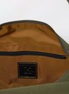 The master-piece x Universal Works Olive Medium Canvas Shoulder Bag is shown from the back as a flat lay against a plain white background. The image highlights the rear of the olive green bag, which features a horizontal webbing strap designed to fit over a suitcase handle. The adjustable and detachable shoulder strap is visible, extending from the sides of the bag.