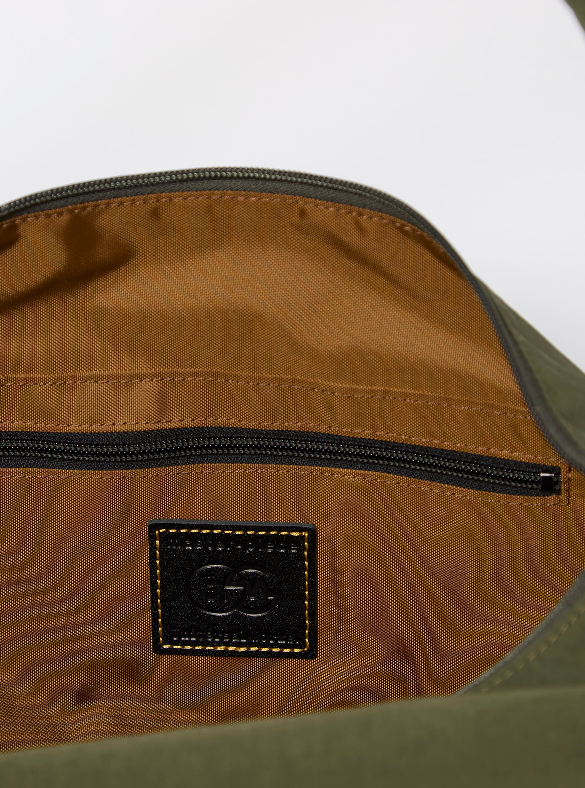 The master-piece x Universal Works Olive Medium Canvas Shoulder Bag is shown from the back as a flat lay against a plain white background. The image highlights the rear of the olive green bag, which features a horizontal webbing strap designed to fit over a suitcase handle. The adjustable and detachable shoulder strap is visible, extending from the sides of the bag.