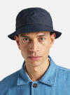 A model wears the Navy Recycled Polytech Beach Hat against a plain, light grey background. The hat is a bucket-style design with a wide, downward-sloping brim and a flat top. The dark navy fabric has a smooth, technical finish with visible tonal stitching around the brim and crown. The model is shown from the head up, with the hat pulled low over the forehead.