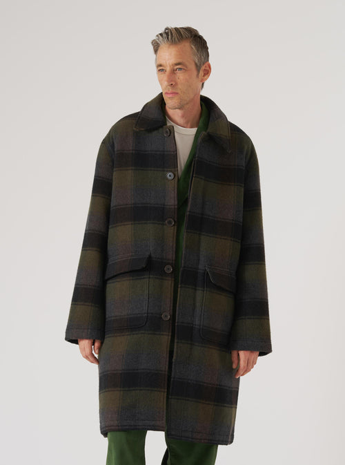 A model poses in a Black/Brown Berlin Wool Check Account Coat against a plain white backdrop. The coat features a black and brown check pattern and a relaxed fit.