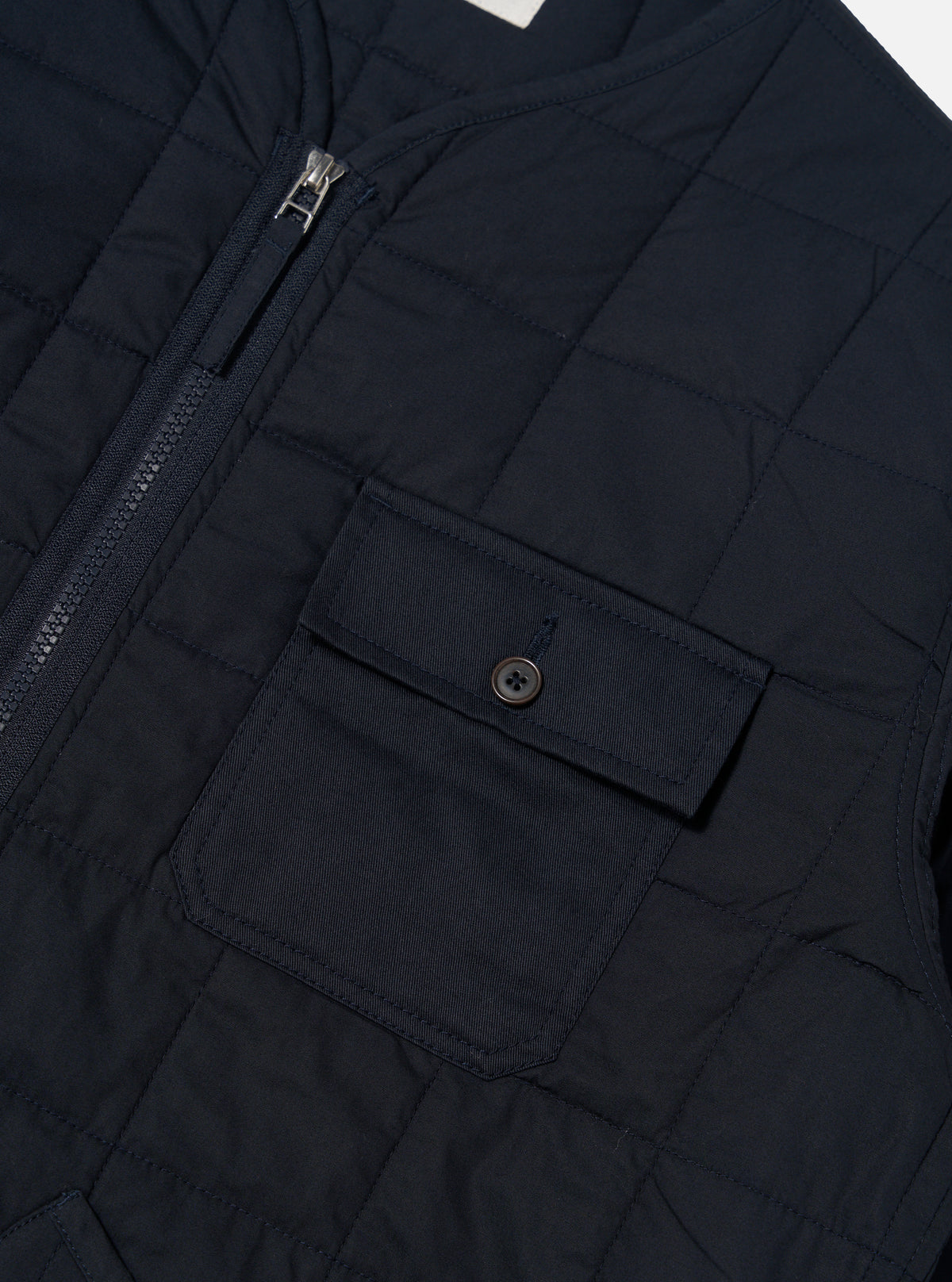 Close-up of the navy quilted cotton military liner jacket, showcasing the zipper, pocket with button, and quilted pattern. The jacket is displayed on a flat surface.