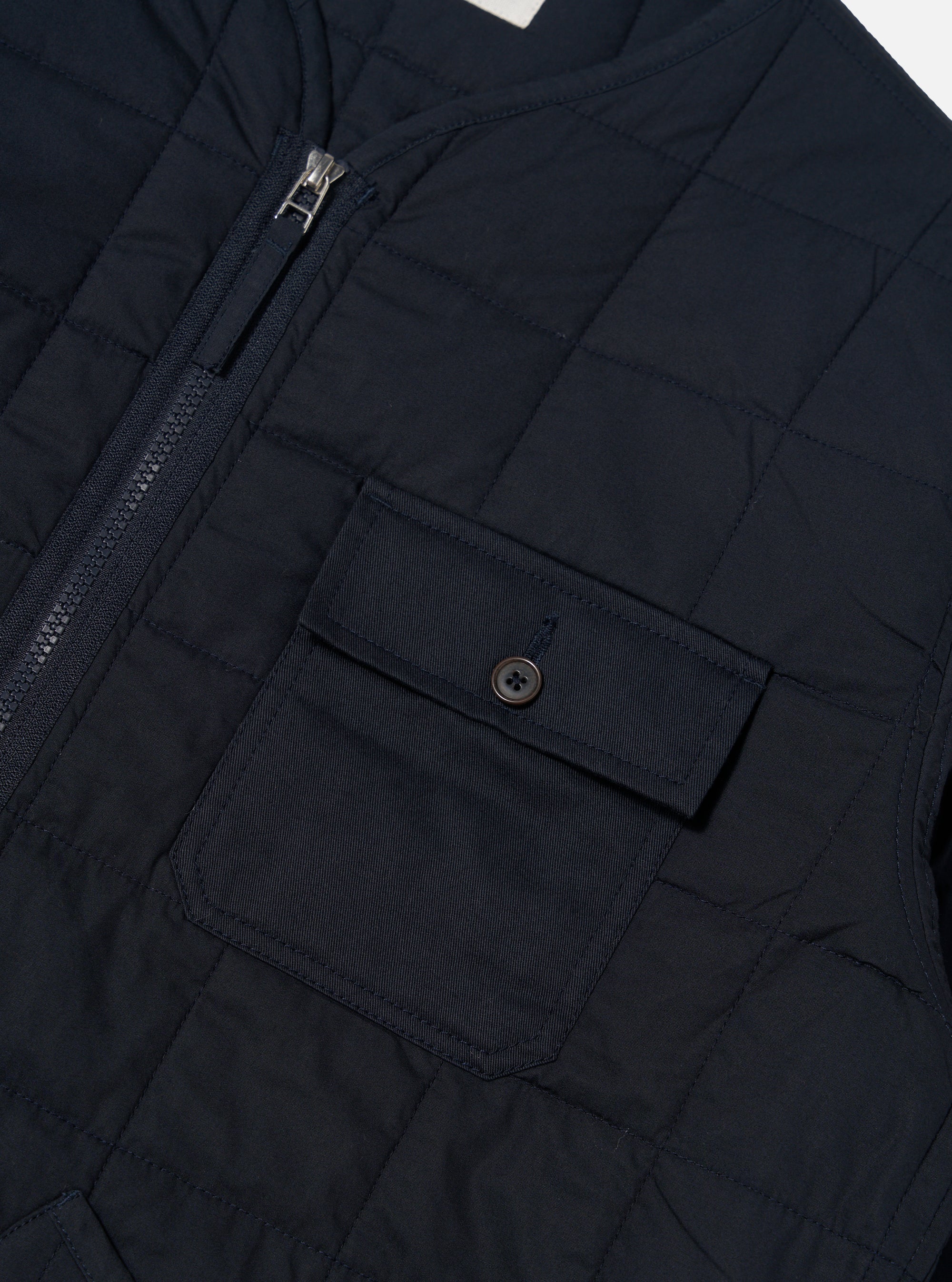 Close-up of the navy quilted cotton military liner jacket, showcasing the zipper, pocket with button, and quilted pattern. The jacket is displayed on a flat surface.