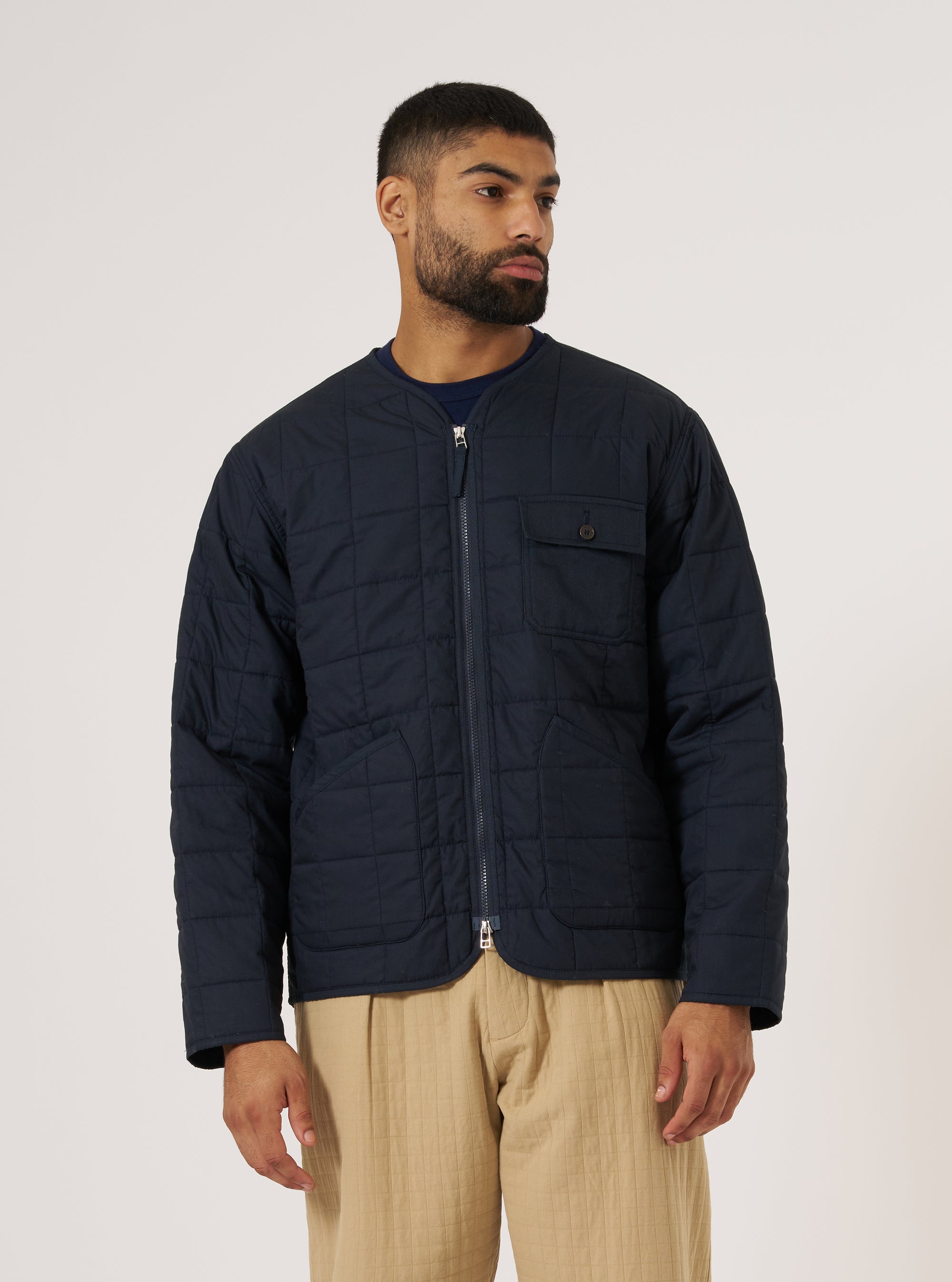 A man poses in a Navy Quilt Cotton Military Liner Jacket against a white backdrop. The navy jacket is paired with tan pants. The jacket features a zip front and quilted pattern.