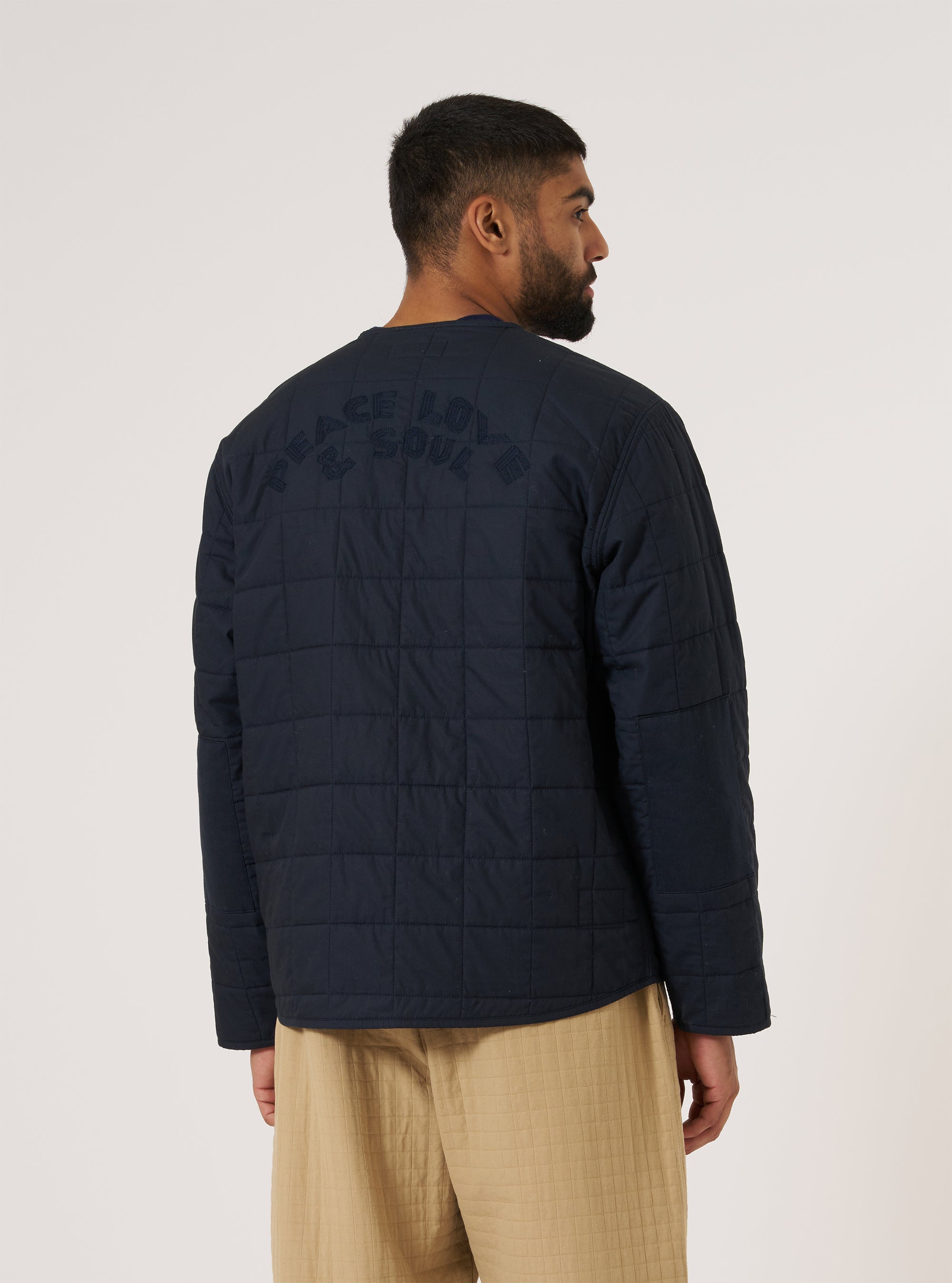 A man stands against a white backdrop, showcasing the back of a navy quilt cotton military liner jacket. The jacket features 'PEACE LOVE & SOUL' embroidered on the upper back.