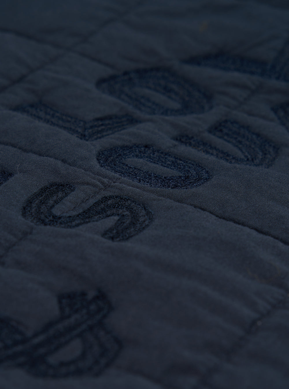 Close-up of the navy quilted cotton Military Liner Jacket, showing the texture and stitching detail on a flat surface. The jacket is a deep navy color.