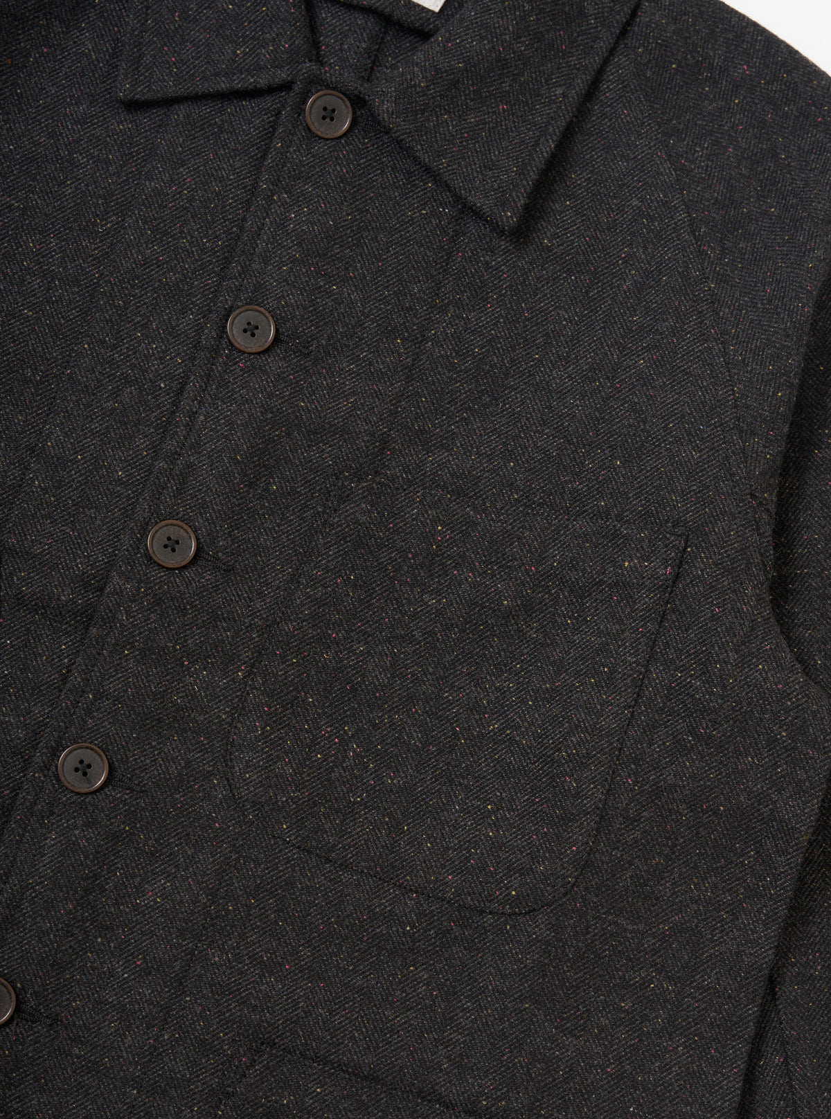 Close-up of the Black Salt N Pepper Tweed Station Jacket. The tweed is dark with flecks of color. Features brown buttons and a pocket. Shot on a white background.