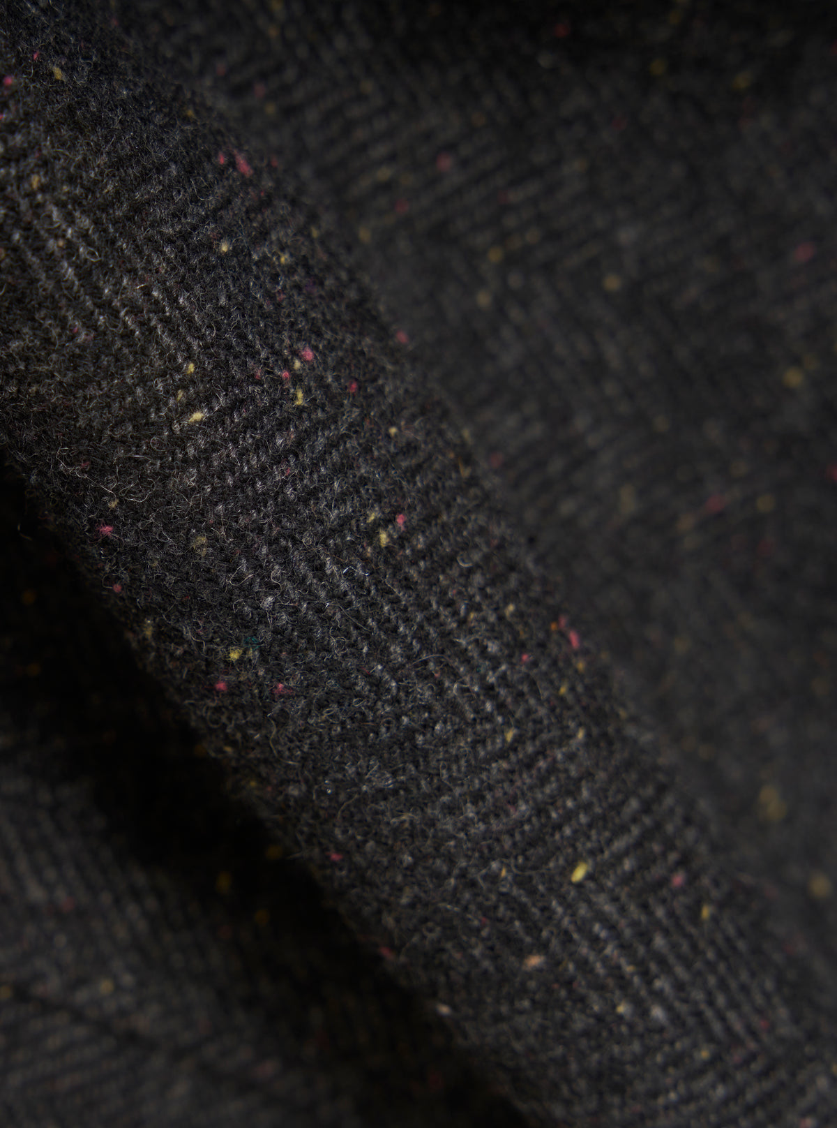 Close-up of the Black Salt N Pepper Tweed Station Jacket fabric. The tweed is a mix of black, salt, and pepper colors with subtle flecks of red and yellow. The fabric is draped to show texture.