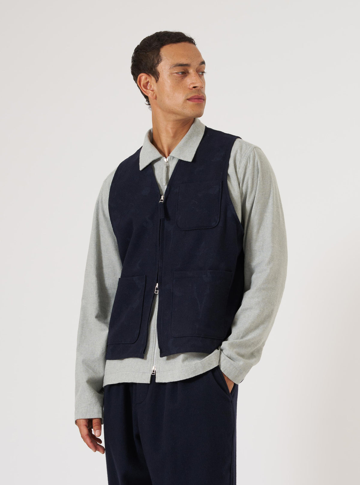 A man poses in front of a white background wearing the Navy Flower Print Flannel Operators Gilet. The gilet is navy with a subtle flower print and a zip front.