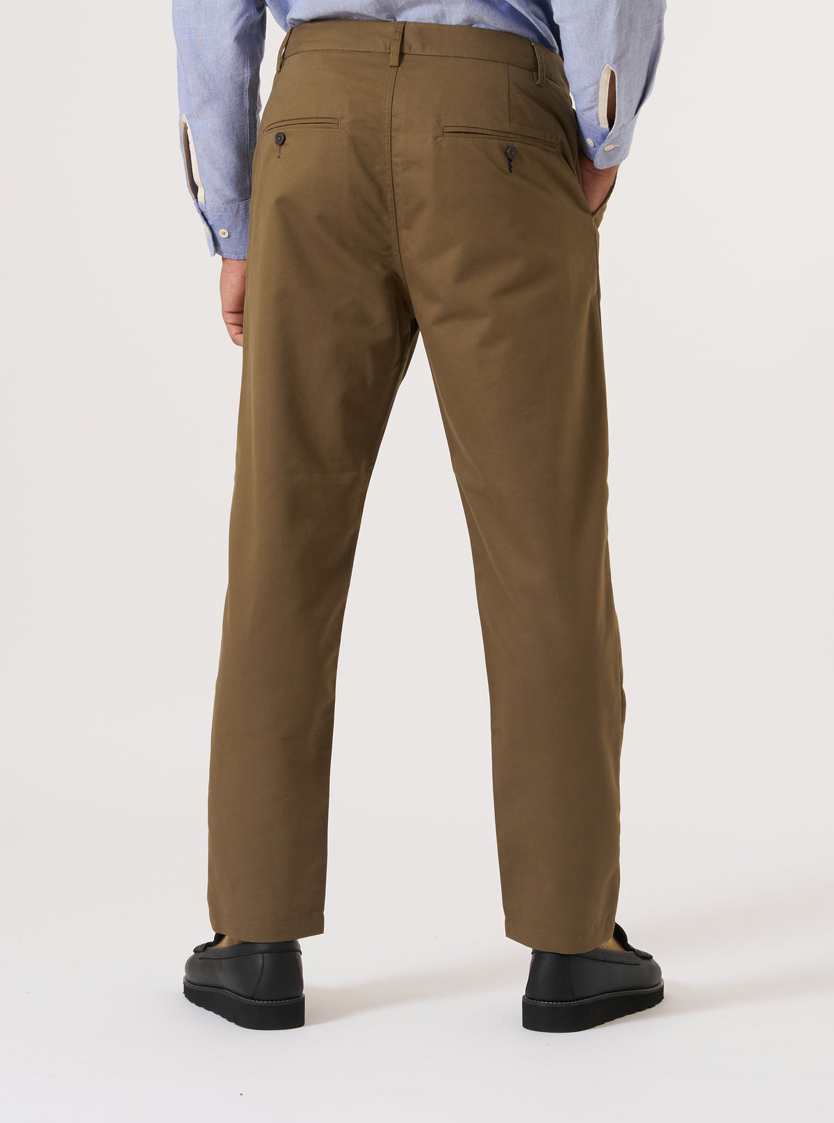 Back view of a model wearing Khaki Brushed Polytech Military Chino pants in a studio setting. The khaki chinos are paired with a blue shirt and black shoes against a white backdrop.