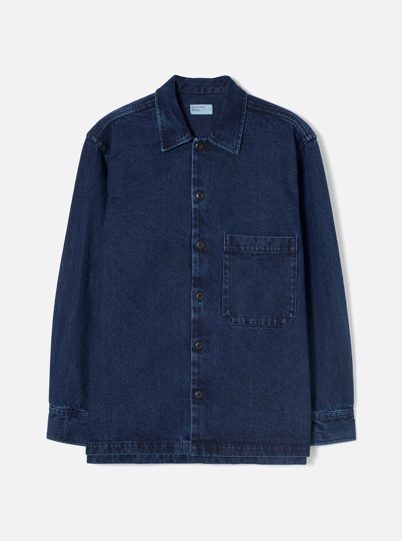 Indigo denim fatigue shirt laid flat on a plain white studio backdrop. Features a pointed collar, full-length button placket with six dark buttons, and a single chest patch pocket on the right. Long sleeves with button cuffs and a straight, slightly layered hem. Woven label visible at the inner collar. Twill weave texture throughout.