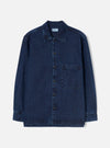 Indigo denim fatigue shirt laid flat on a plain white studio backdrop. Features a pointed collar, full-length button placket with six dark buttons, and a single chest patch pocket on the right. Long sleeves with button cuffs and a straight, slightly layered hem. Woven label visible at the inner collar. Twill weave texture throughout.