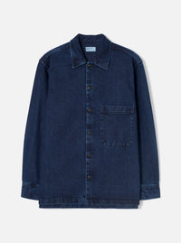 Indigo denim fatigue shirt laid flat on a plain white studio backdrop. Features a pointed collar, full-length button placket with six dark buttons, and a single chest patch pocket on the right. Long sleeves with button cuffs and a straight, slightly layered hem. Woven label visible at the inner collar. Twill weave texture throughout.