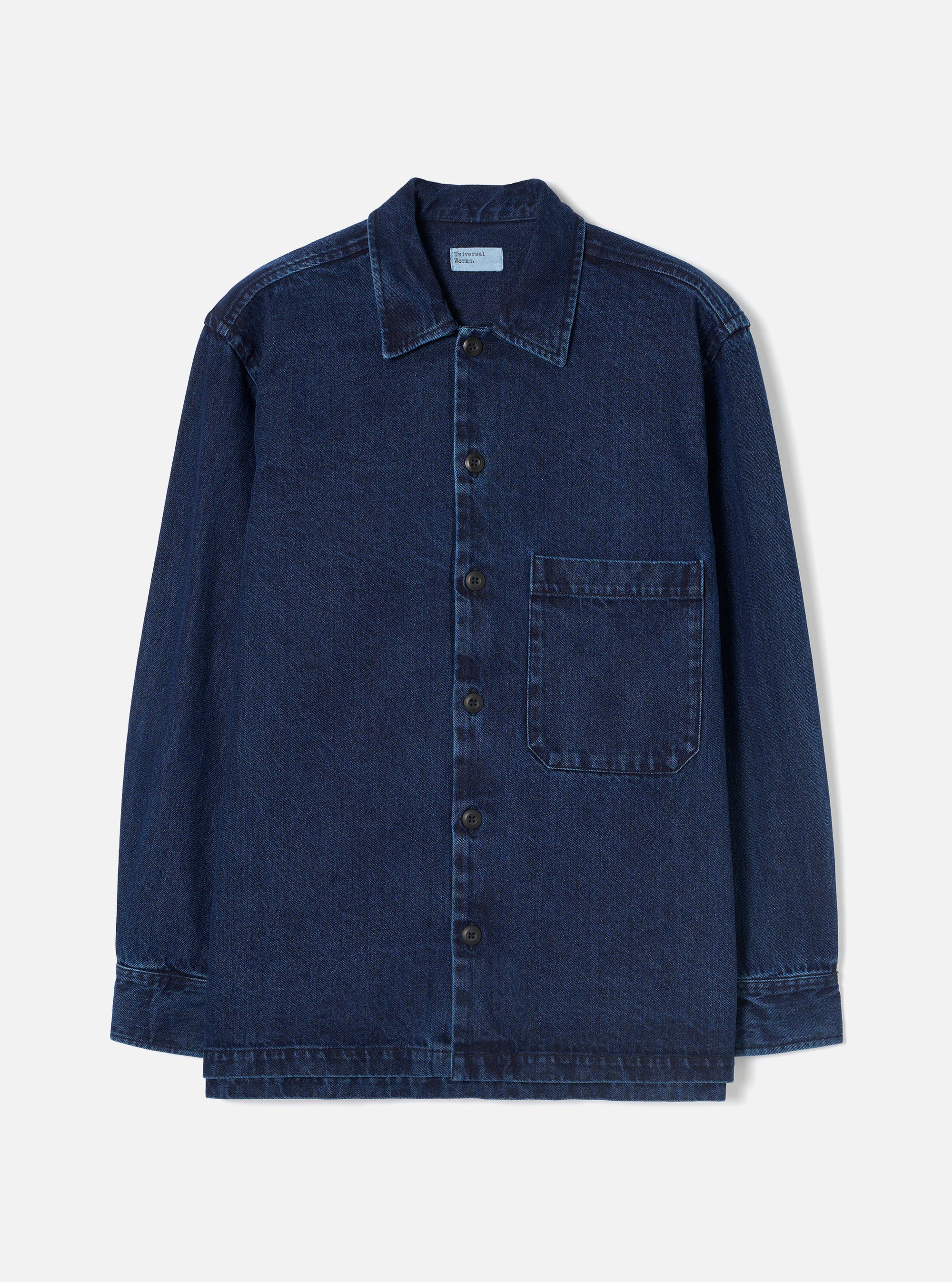 Indigo denim fatigue shirt laid flat on a plain white studio backdrop. Features a pointed collar, full-length button placket with six dark buttons, and a single chest patch pocket on the right. Long sleeves with button cuffs and a straight, slightly layered hem. Woven label visible at the inner collar. Twill weave texture throughout.