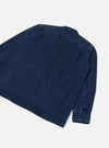 Rear flat-lay of an indigo work denim fatigue shirt on a light grey backdrop. The shot shows the back panel with topstitched seams across the shoulders and a single back yoke. One long sleeve is visible, ending in a button cuff with two dark buttons. The denim fabric has a medium-weight woven texture.