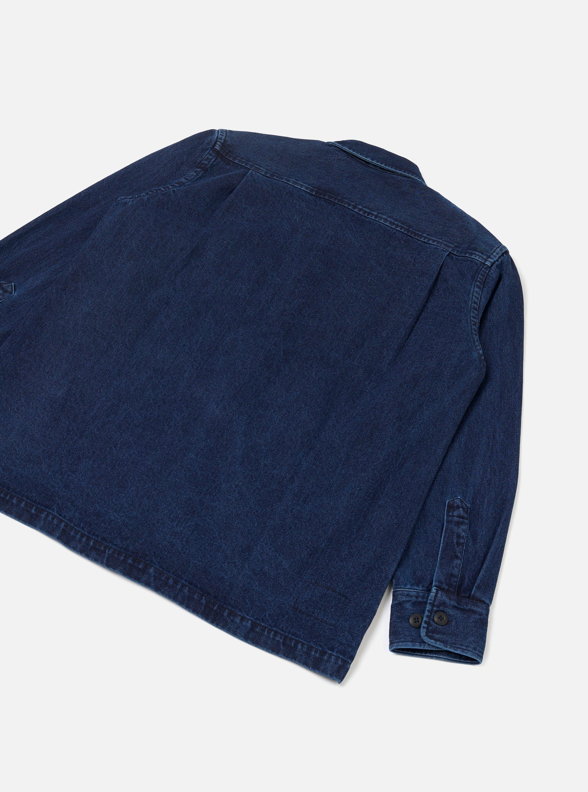 Rear flat-lay of an indigo work denim fatigue shirt on a light grey backdrop. The shot shows the back panel with topstitched seams across the shoulders and a single back yoke. One long sleeve is visible, ending in a button cuff with two dark buttons. The denim fabric has a medium-weight woven texture.