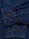 Close-up of the cuffs on an indigo denim fatigue shirt. The twill weave fabric shows deep blue tones with subtle texture. Each buttoned cuff features two dark horn-style buttons with amber thread bartacks. A chest pocket flap with a single button is visible at the top of the frame. No background is visible.