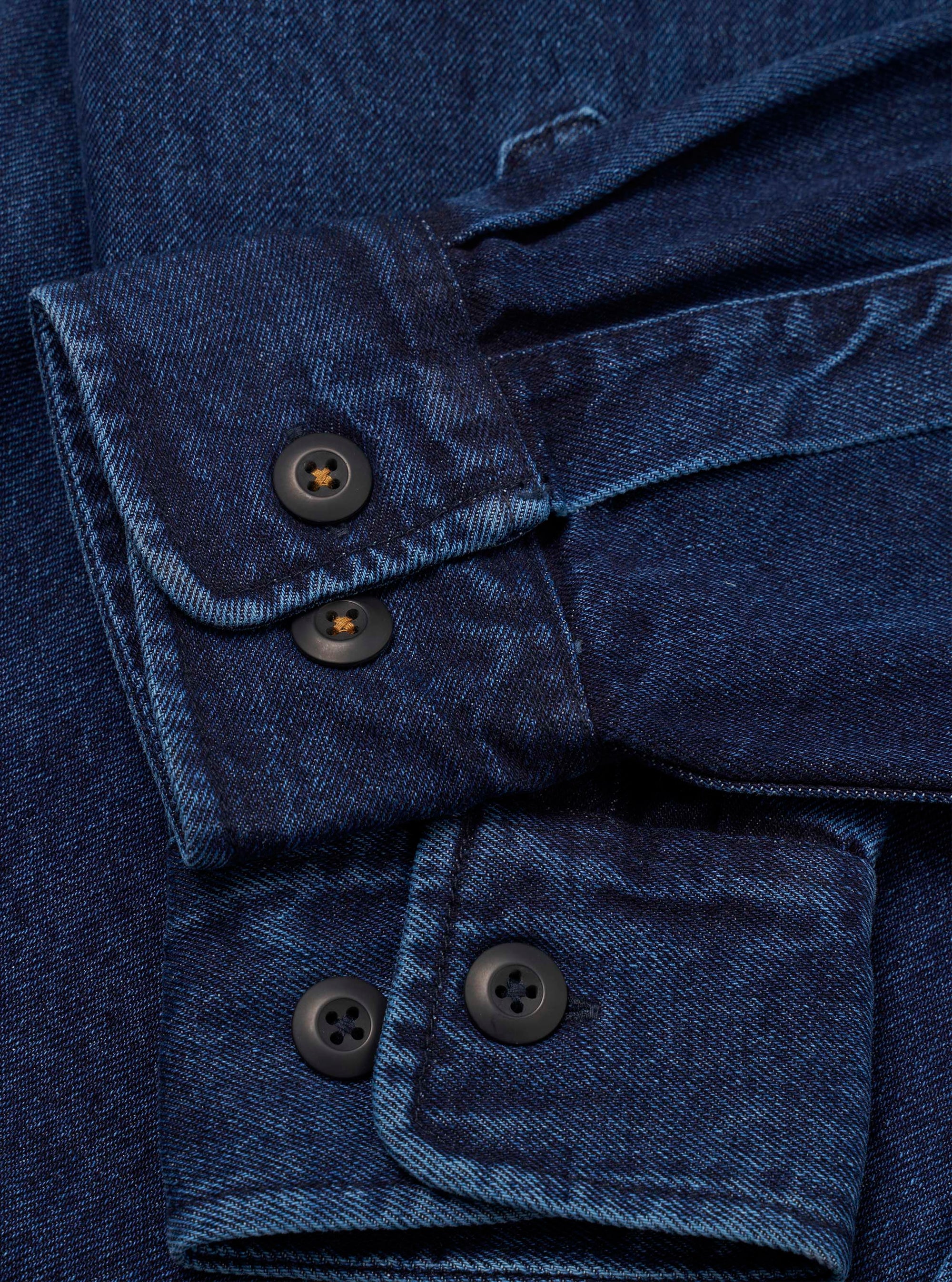 Close-up of the cuffs on an indigo denim fatigue shirt. The twill weave fabric shows deep blue tones with subtle texture. Each buttoned cuff features two dark horn-style buttons with amber thread bartacks. A chest pocket flap with a single button is visible at the top of the frame. No background is visible.