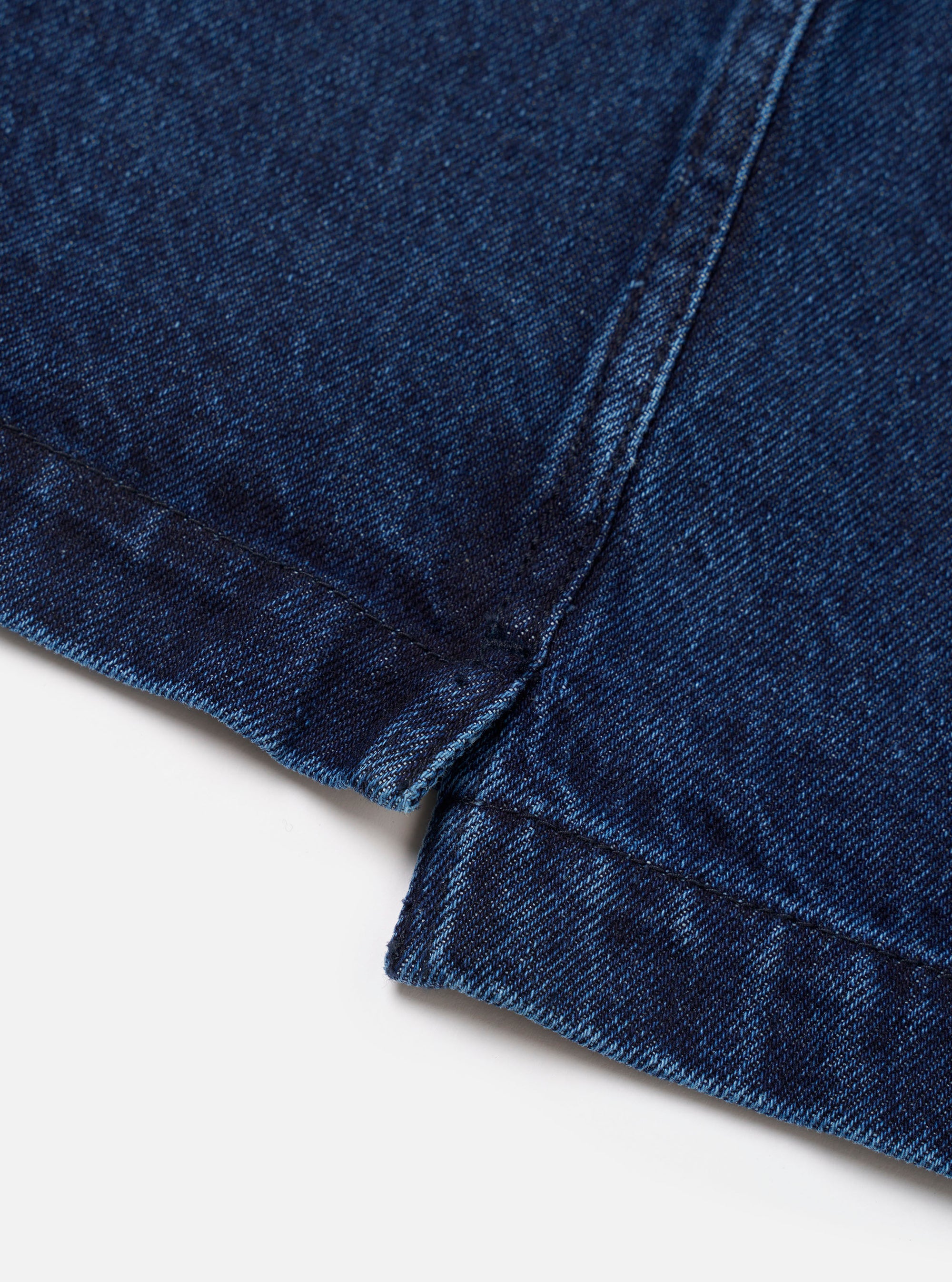 Close-up detail of the indigo work denim fatigue shirt laid flat on a white surface. The shot focuses on the hem and side seam junction, showing the folded and stitched hem with a small side vent. Dark indigo denim fabric with a tight twill weave is visible throughout.