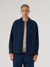 Male model wearing an open indigo denim fatigue shirt over a grey marl crew-neck top and matching indigo denim trousers, against a light grey studio backdrop. The shirt features a spread collar, dark buttons down the front placket, one chest patch pocket, and buttoned cuffs. Shot cropped at mid-thigh, model facing forward.