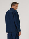 Back view of a model wearing the Indigo Work Denim Fatigue Shirt in dark indigo denim. The rear yoke seam and long sleeves with buttoned cuffs are clearly visible. The shirt is worn untucked over matching indigo denim trousers. Shot from mid-thigh up against a light grey studio backdrop, model looking down.