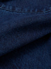 Close-up detail of the indigo work denim fatigue shirt showing the twill weave texture of the dark indigo fabric. A curved chest patch pocket with topstitching is visible at the top. The fabric is folded to reveal depth and weight. No model or mannequin present.