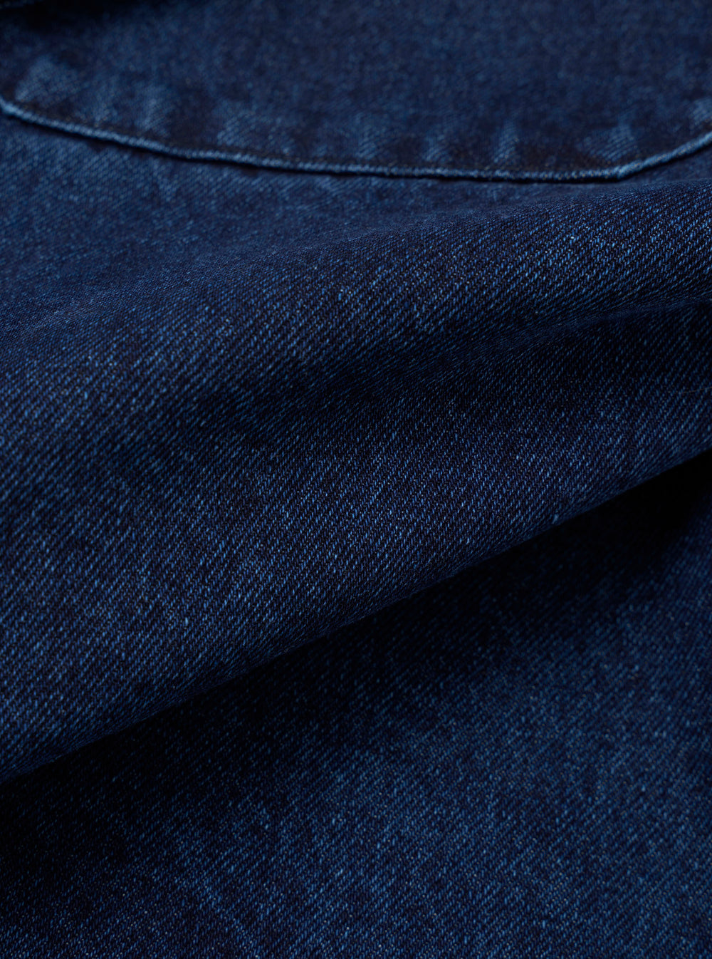 Close-up detail of the indigo work denim fatigue shirt showing the twill weave texture of the dark indigo fabric. A curved chest patch pocket with topstitching is visible at the top. The fabric is folded to reveal depth and weight. No model or mannequin present.