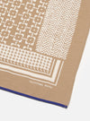 Close-up of the Sand Single Jersey Neckerchief on a white surface. Features a tan fabric with white geometric patterns and a blue hem. 'Universal Works' printed on it.