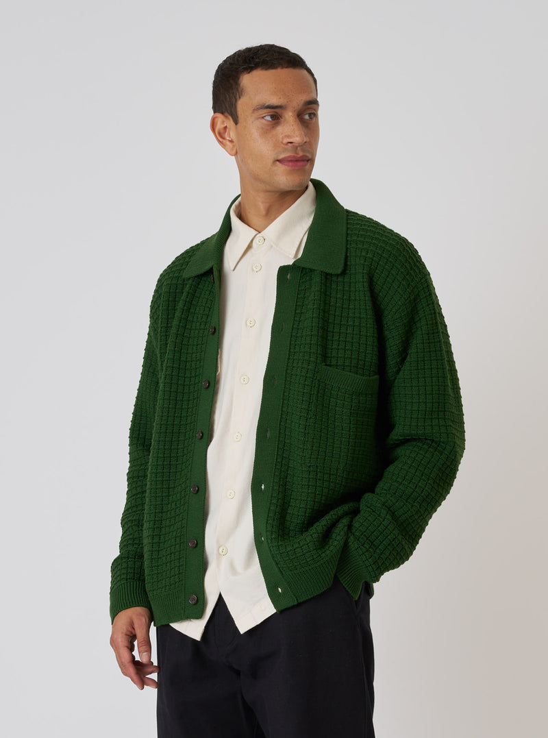 A model poses in front of a white background wearing a Green Textured Merino Knit Charlie Cardigan, a collared shirt, and black pants.