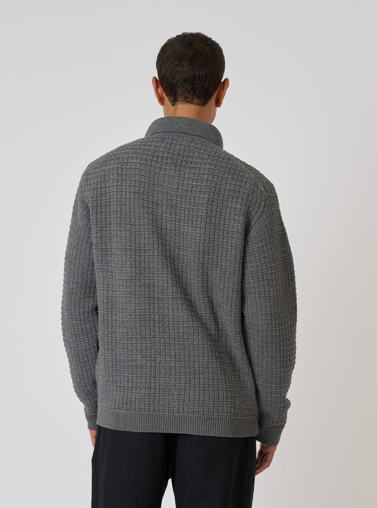 Back view of a model wearing a grey textured merino knit Charlie Cardigan. The cardigan has a waffle knit texture. Studio shot on a white background.