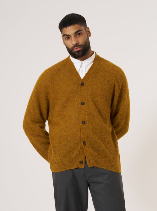 A model poses in front of a white background wearing a Gold Alpaca Knit David Cardigan, a white shirt, and gray pants. The cardigan is buttoned and has a relaxed fit.