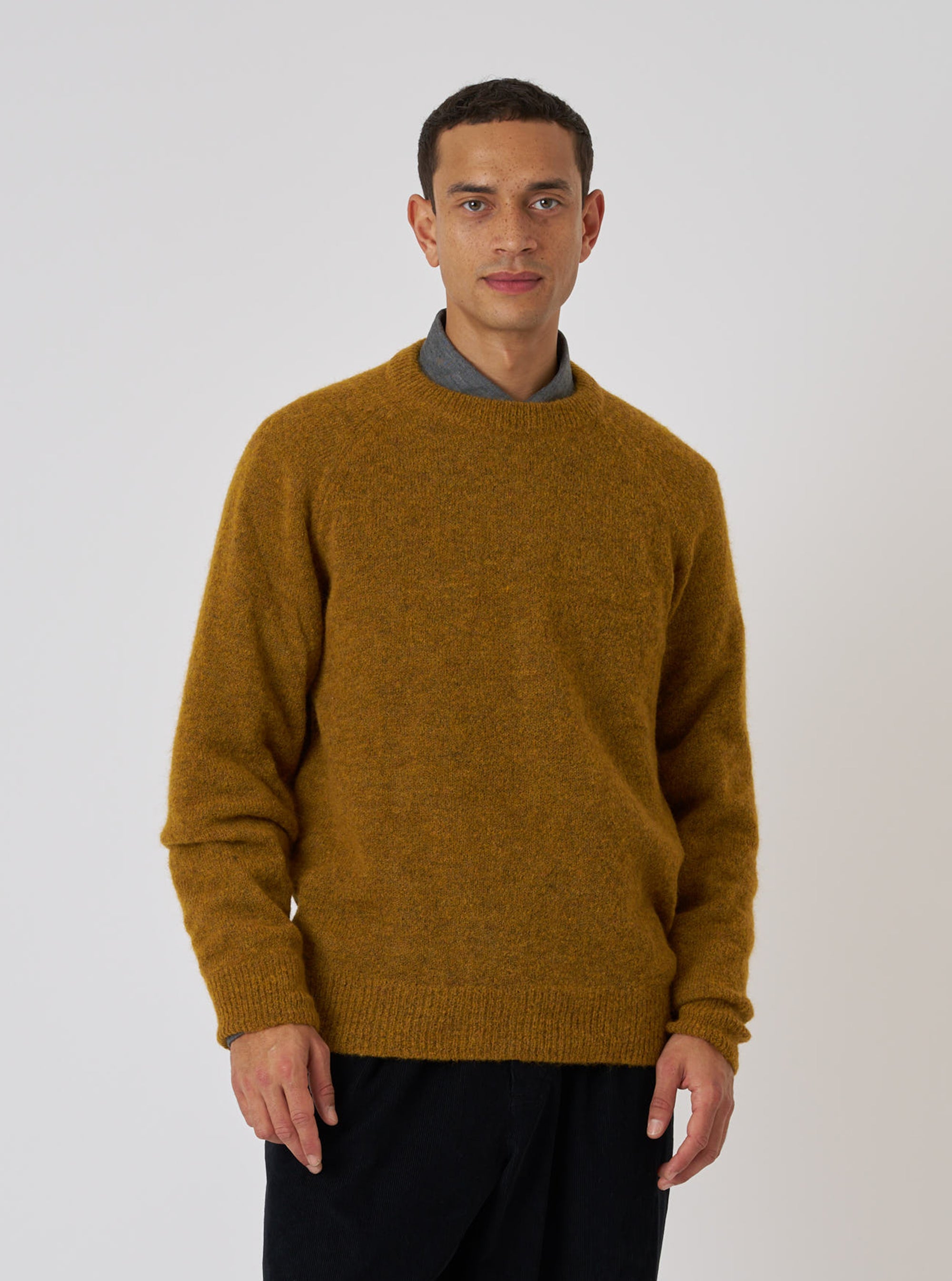 A man stands against a white backdrop, modeling the Gold Alpaca Knit Lyon Crew. The gold sweater is paired with dark pants and a gray shirt.