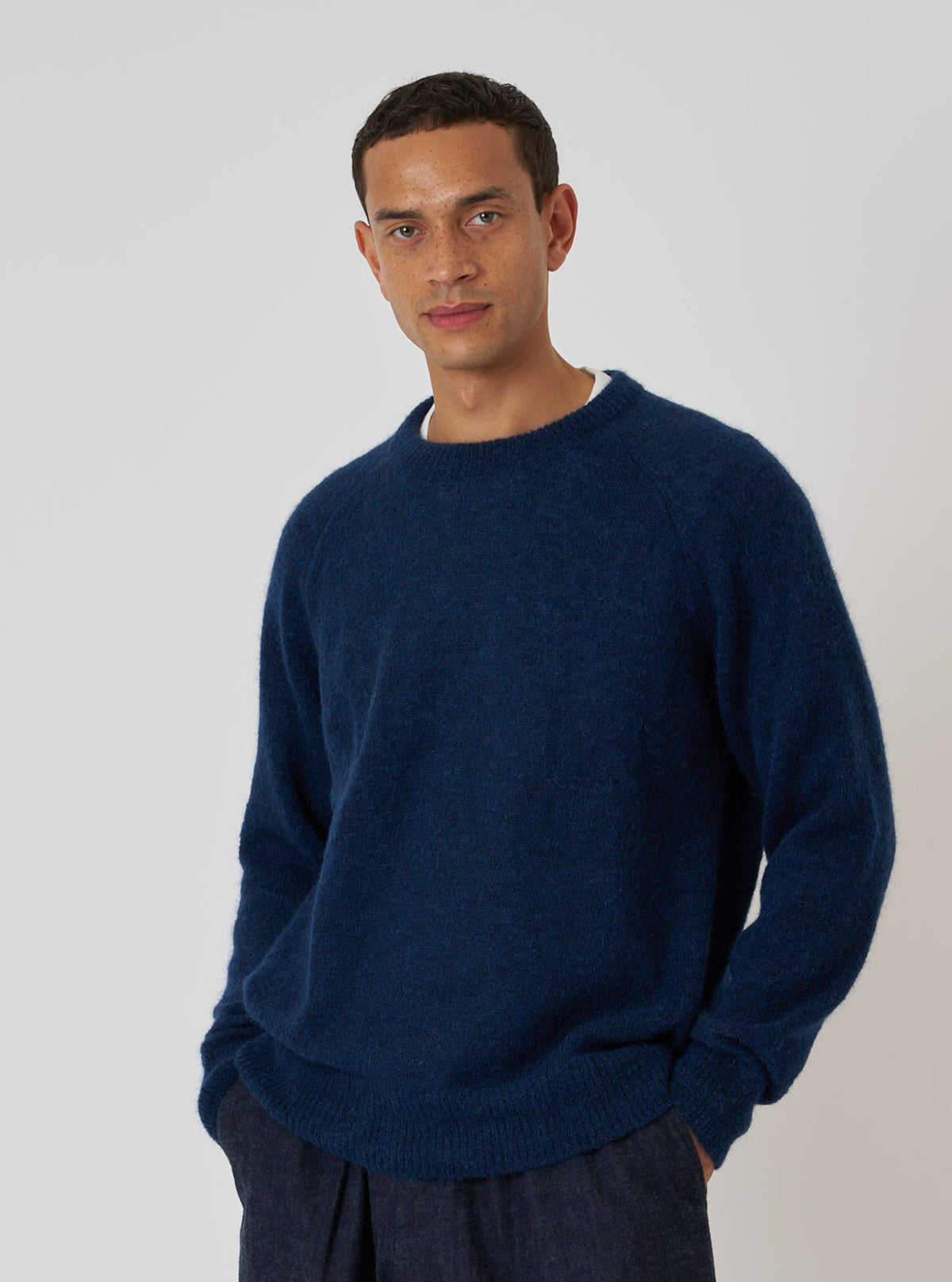 A man poses in front of a white backdrop, showcasing the Navy Alpaca Knit Lyon Crew. The sweater is a rich navy blue, and he wears dark trousers.