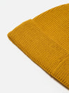 Close-up of a gold-colored, ribbed knit Gold Merino Knit Watch Cap on a white surface. The cap is folded, showing the texture of the merino wool.