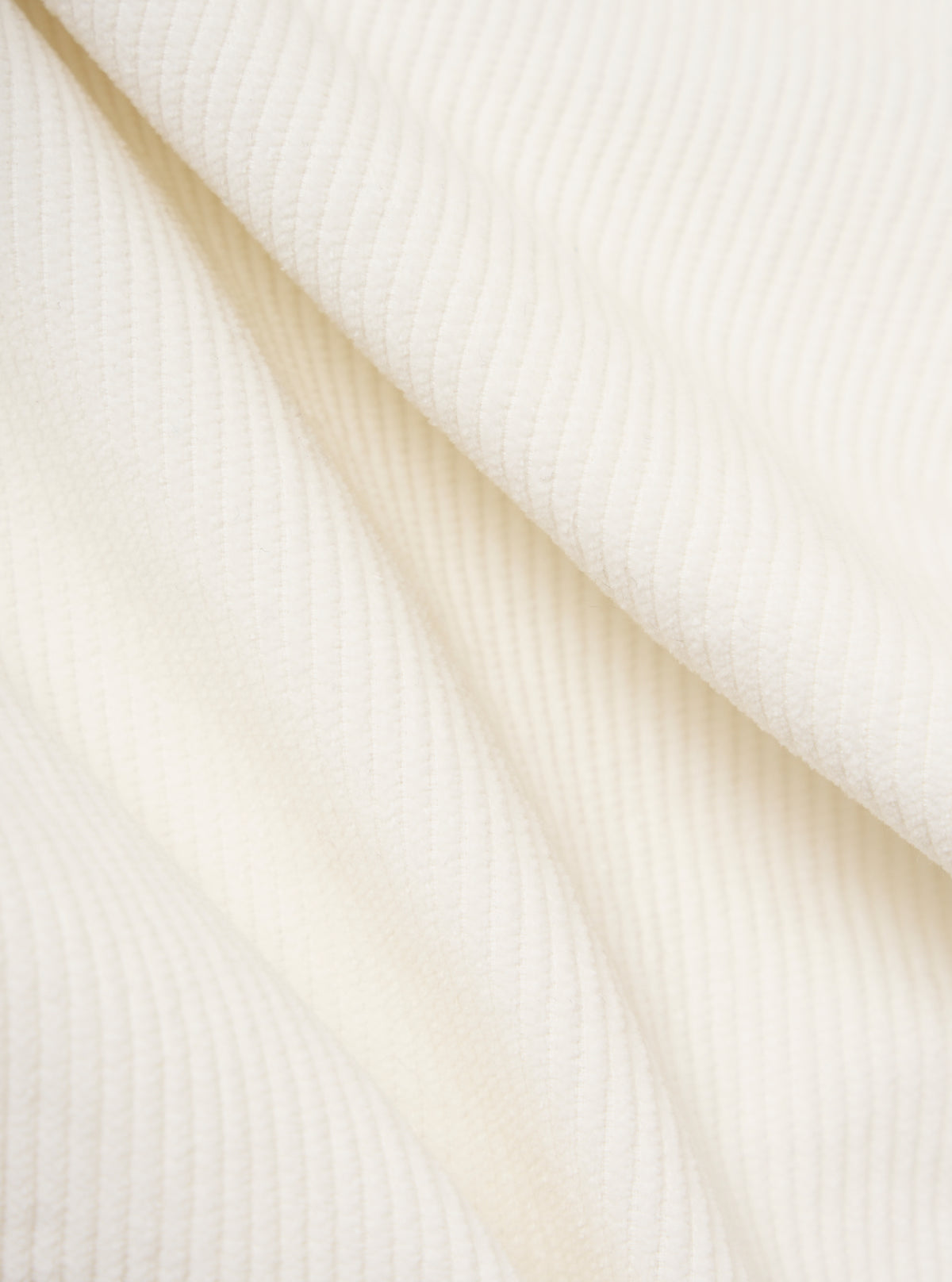 Close-up of the Ecru Cord Pleated Track Pant fabric. The soft, textured corduroy material is draped on a plain white surface, highlighting its subtle pleats and creamy ecru color.