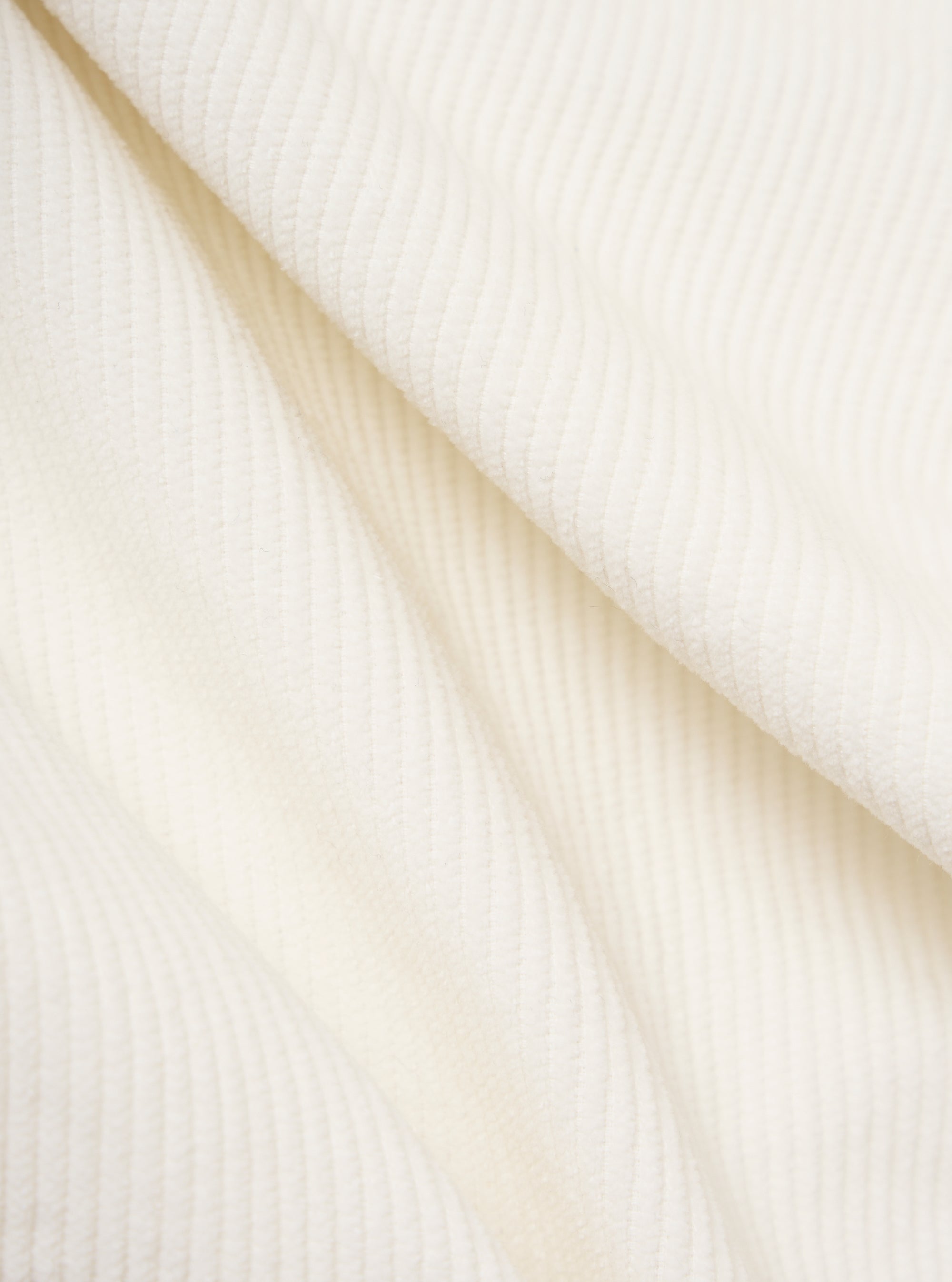 Close-up of the Ecru Cord Pleated Track Pant fabric. The soft, textured corduroy material is draped on a plain white surface, highlighting its subtle pleats and creamy ecru color.