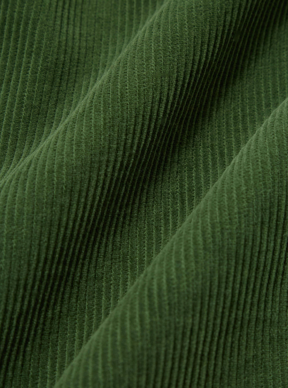 Close-up of the green corduroy fabric of the Green Cord Pleated Track Pant, showing the texture and pleats. The fabric is draped on a flat surface.