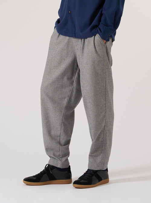 Model wears Grey Leon Tweed Pleated Track Pant in a studio shot. The grey tweed pants have a relaxed fit. Worn with black sneakers. Clean white background.