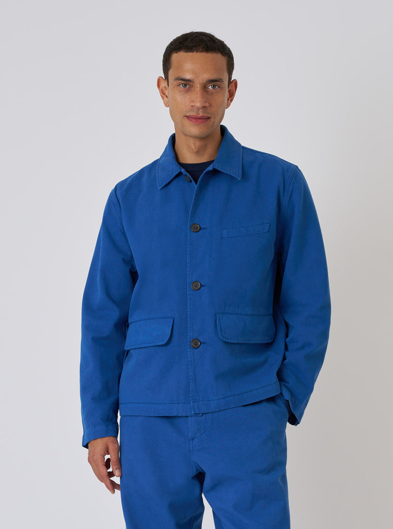 A model poses in a Navy Derek Twill Warmus Jacket and matching pants against a white backdrop. The jacket is a vibrant blue twill with button closure and patch pockets.
